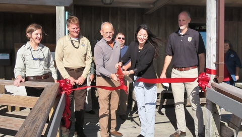 A group of five men and women cutting a ribbon with a pair of oversized scissors. There are a few onlookers in the background.