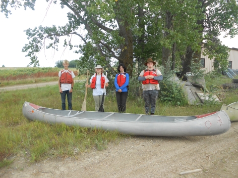 4 youth standing in a line behind an aluminum canoe outside