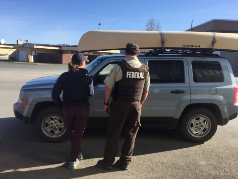 two individuals (one wearing a federal law enforcement vest) talking to a driver in a stopped car that has a boat attached to the roof 