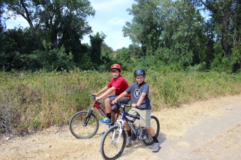 Two children sit on bikes on a trail in front of shrubs and trees