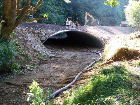 Dry streambed with new culvert for fish passage installed, with heavy machinery on the road above