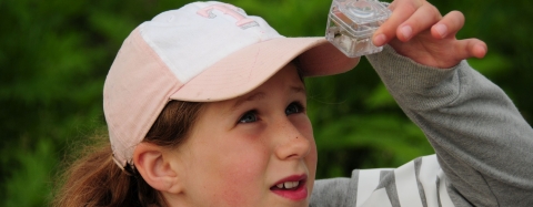 A girl in a pink cap looks up into a small clear bug box that she is holding.