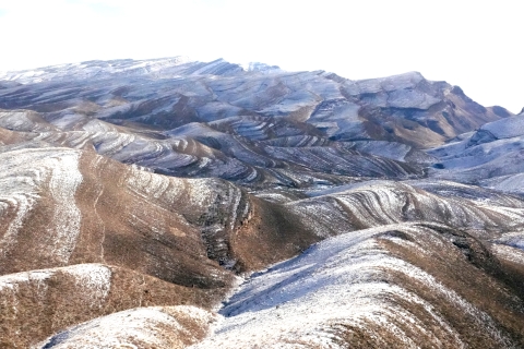 Snow covers areas of a mountain range. The mountains have short, brown vegetation growing on them. This makes the white snow contrast against the brown vegetation, and the mountains appear to have waves of white and brown across them. The mountains are closer to the viewer at the bottom of the photo and get farther away moving towards the top of the photo, until there is a white sky at the top.