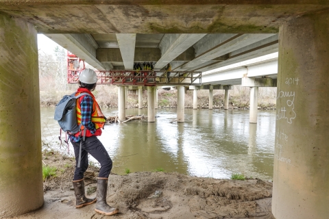 Person standing on river bank beneath bridge watching work crew on catwalk working below the bridge