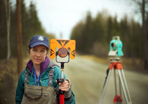 Woman holding surveying equipment stands on a rural Alaskan dirt road