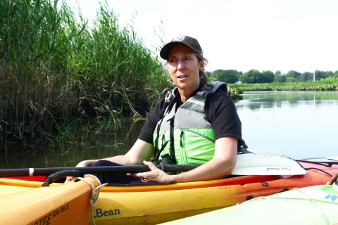 woman in lifevest in kayak in water near a shore with tall grasses