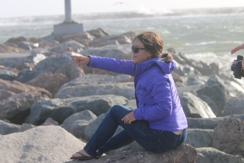 A woman in a purple jacket pointing at something off screen while sitting on a pile of rocks