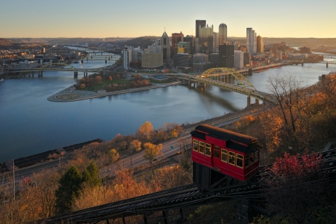 a city skyline photo taken from a hillside. Skyscrapers surrounded by a river, and multiple bridges reaching across the water. A funicular car can be seen in the foreground, as well as trees lining a street. It appears to be autumn due to the vibrantly colored trees.