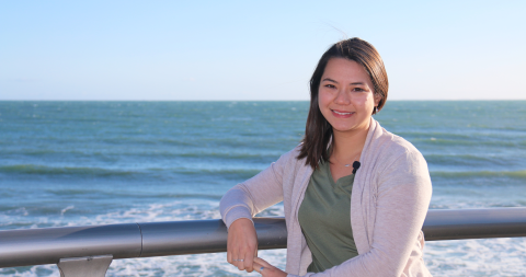 A woman smiling at the camera and leaning against a railing that overlooks the ocean