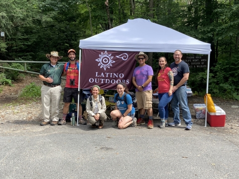a group of people stand by an outdoor tent. A flag raised on the tent says "latino Outdoors" with a symbol of a sun above it. THe people are smiling wearing hats, binoculars and backpacks