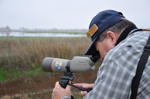 A man in a cap peers through a scope aimed at tall grass around a lake 