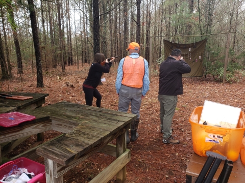  A man and a women holding firearms face the woods with a man with a bright orange vest on in between them.
