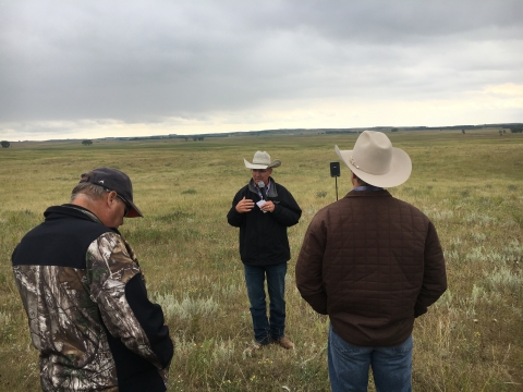 A man wearing a cowboy hat speaks to a small group of people on a grassland