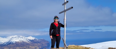 A woman wearing a hat and sunglasses on a high summit overlooking snowcapped mountains and ocean. She stands next to a metal structure.