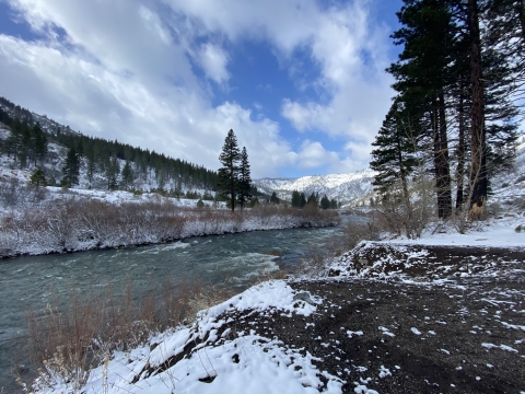 A landscape photo of a river flowing between mountains dusted with snow under a partly cloudy blue sky.