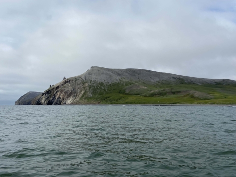 Crowbill Point, covered in green and grey with grass and dirt, stretches into the blue waves of the Chukchi Sea.