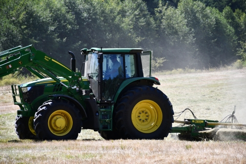 A green and yellow tractor tows a mowing attachment across a dusty field of grass with evergreens in the background.