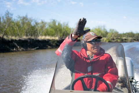 Close up of Chris Tulik operating a boat on a river while waving his hand at someone outside of the image. 