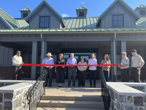 6 people behind red ribbon in front of Tan building with green roof, 2 others hold ribbon, another watches