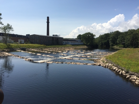 A river with rocks placed in lines flows by an old brick mill complex