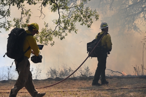 A cameraman follows a firefighter who is walking toward small flames and a smoky forested landscape. The firefighter has a hose slung over his shoulder and is dragging the rest on the ground.
