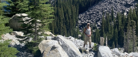 A man stands on the side of a rocky mountain with rocks, trees and greenery around him.