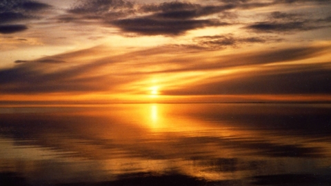 An orange sunset over calm water. The sky is scattered with clouds and the water is reflecting the sun and sky.