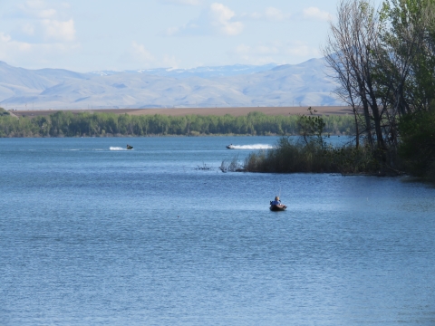 A kayaker is fishing and two people are riding personal watercraft on a calm lake. There are trees on a point in the foreground and on the distant shoreline, as well as mountains in the distance. 
