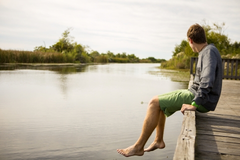 A man sitting on the edge of a dock with his legs dangling over the river. He's looking up river away from the camera