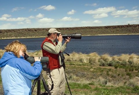 Two people stand near a body of water and look at wildlife through lenses-- one a camera lens, the other a scope.