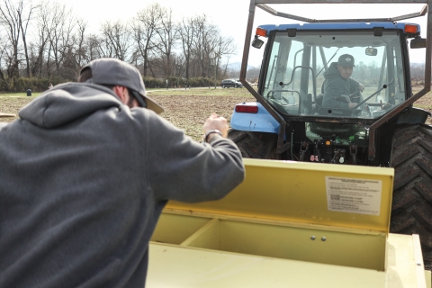 One person looks in a seed drill while another watches from a tractor cab
