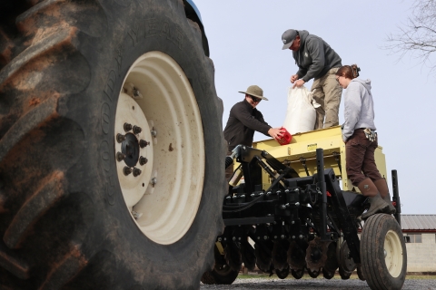 Three people standing on a tractor-pulled seed planter, with a large tractor tire in the foreground.