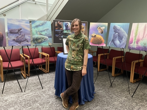 an artist, Cal Robinson, poses with a smile in front of a display of canvas prints of their digital paintings for the endangered species act 50th anniversary. Species include kemps ridley sea turtles, atlantic salmon, rusty patched bumble bee, manatee, and ka'u silversword