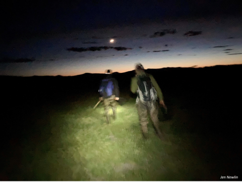 researchers walking into Klamath Marsh National Wildlife Refuge at dusk. The picture is dark. The sun is setting in the distance and you can see the moon in the sky. Two people with their backs to the camera are walking into the refuge.