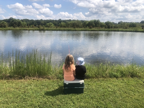 Two kids sitting on a cooler fishing in a pond on a sunny day.