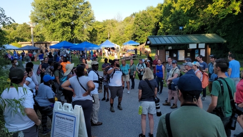 A crowd of people stand together outside with tents and small buildings in the background