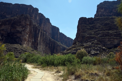 A wooden walk way passes through desert riparian vegetation in the foreground, with canyon cliffs in the background.