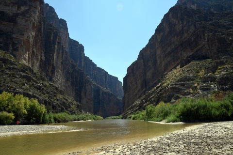A sandy beach and river in the foreground, with desert riparian plants and tall rocky canyon cliffs in the background.