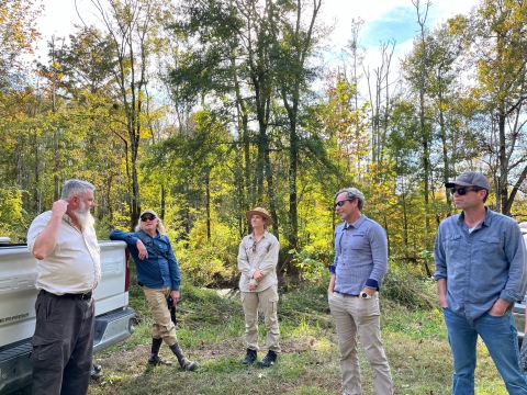 Five people stand a few feet apart from one another in a wooded area to discuss water management challenges.