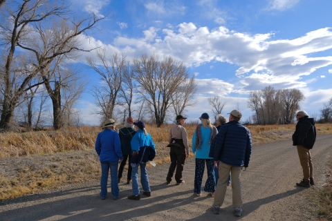 Suzanne Beauchaine and locals gather in a group on a gravel trail during the wildlife walk. cottonwood trees and cloudy, blue skies in the background.