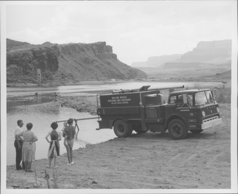 Fish hatchery truck, backed up to a body of water, with a pipe running from a tank in the bed of the truck, down to just above the surface of the water. Four (4) people watch from the left foreground.