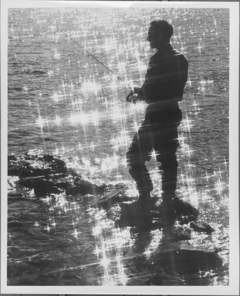 Man, standing on a narrow rock formation in a body of water, fishing with a fishing rod. The sunlight sparkles on the water. 