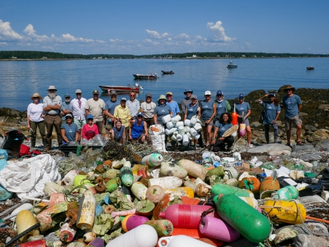 Two dozen or so people stand and kneel near a big pile of plastic debris they collected and removed from a Maine coastal island.