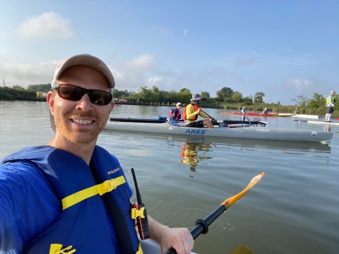 A man wearing a blue life-preserver, blue shirt sunglasses and baseball cap smiles toward the camera from a kayak on Chesapeake Bay. His kayak isn't visible, but his paddle is. Other kayakers are visible on the greenish water behind him.