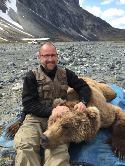 A biologist sits next to a sedated brown bear on a gravel plain in the mountains.