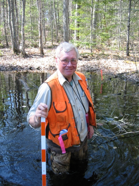 A man holding a yard stick and wearing an orange vest and waders standing in a shallow pond in a forested area