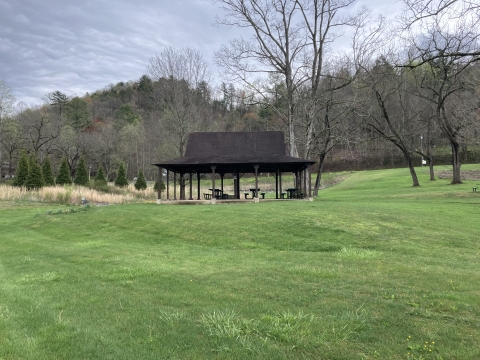 A photo of a picnic shelter with a steep pitched roof standing in a grassy field. 