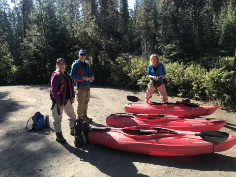 Three people geared up for frog surveys stand next to three red kayaks outdoors, on gravel with trees in the background.