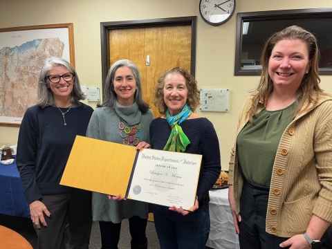 Four women standing in a row, smile at camera. Two in center are holding Bridget's award certificate.