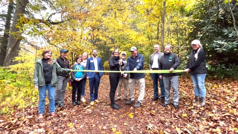 A group of people stand in front of a forest holding a ribbon while a woman cuts the ribbon with a huge pair of scissors. 